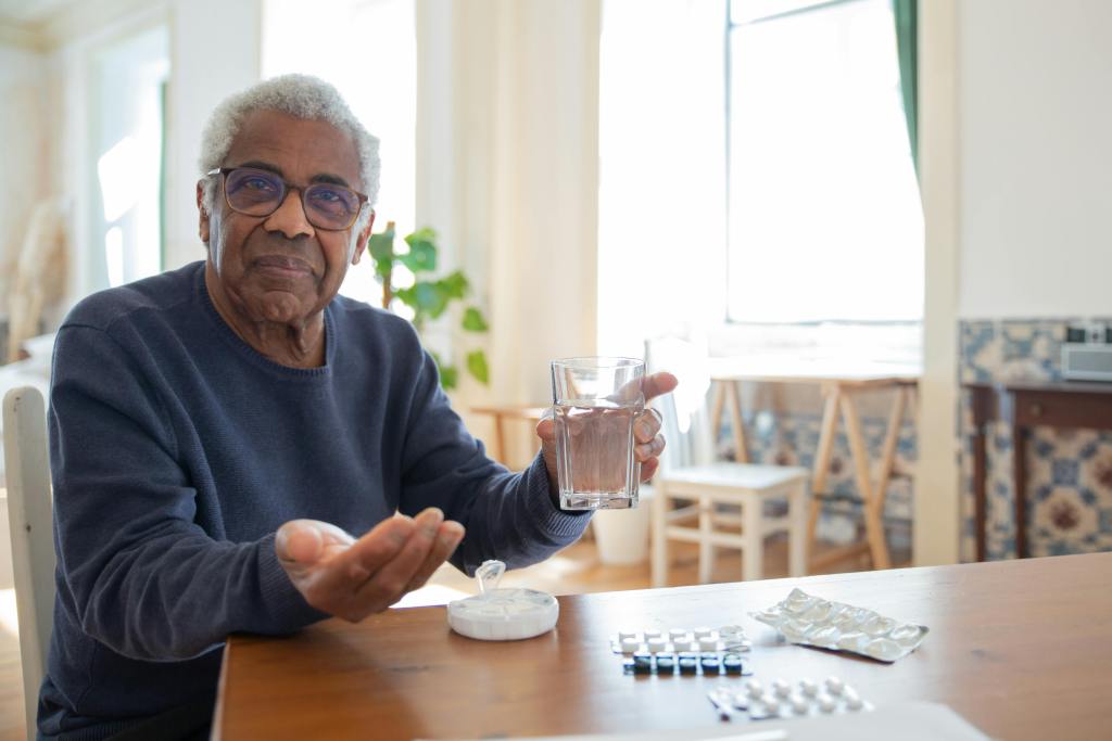 Elderly man sitting at a table at home, holding a glass of water and medication, emphasizing healthcare routine.