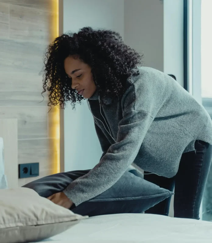 A woman making a bed in a modern bedroom with natural light, reflecting a cozy and tidy home environment.