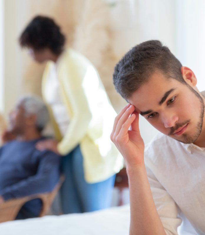 Young man in thoughtful pose indoors with family members in the background, expressing contemplation and care.